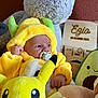 baby, plush_toy, pikachu, costume, pacifier, teddy_bear, avocado_plush, yellow_clothing, child, soft_toys, infant, cute, indoor, portrait, smile, hand, face, wooden_sign, decor, cuddle