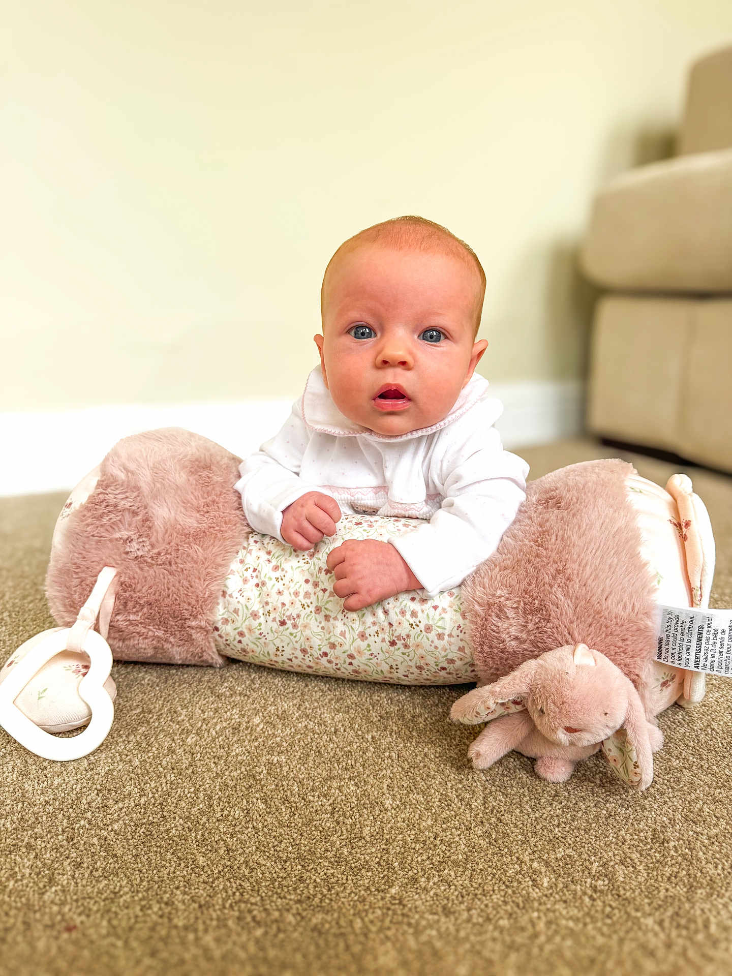 Larna joined the competition — help win amazing prizes! baby, infant, child, carpet, plush_pillow, soft_toy, floor, indoor, blue_eyes, white_clothing, curious_expression, hands, couch, blurred_background, home, cozy, face, portrait, cute, toddler