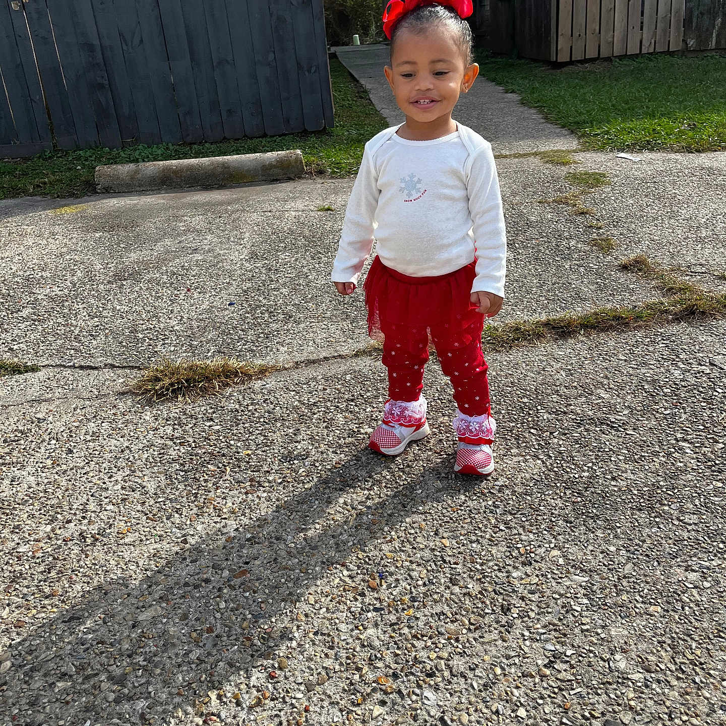 Emmy Levangie joined the competition — help win amazing prizes! building, child, concrete, daylight, fence, grass, happy, outdoor, person, playful, red_bow, red_clothing, shadow, shoes, sidewalk, smiling, standing, sunny, toddler, white_shirt