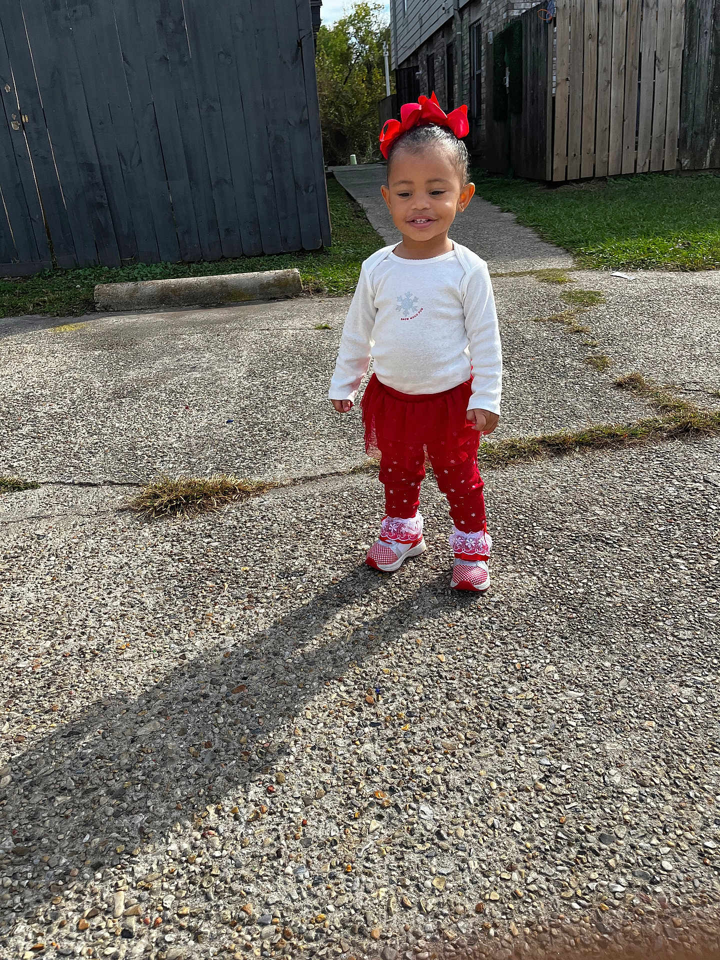 Emmy Levangie joined the competition — help win amazing prizes! toddler, child, outdoor, concrete, red_bow, red_clothing, white_shirt, shadow, grass, fence, sidewalk, smiling, standing, shoes, sunny, daylight, building, happy, person, playful