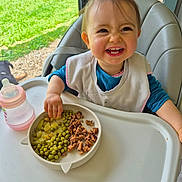 élyne a rejoint le concours — aidez-le/la à gagner de superbes lots ! toddler, child, high_chair, plate, peas, meat, pickles, baby_bottle, smiling, happy, indoor, feeding, food, table, window, natural_light, clothing, bib, hand, face