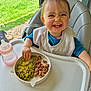 toddler, child, high_chair, plate, peas, meat, pickles, baby_bottle, smiling, happy, indoor, feeding, food, table, window, natural_light, clothing, bib, hand, face