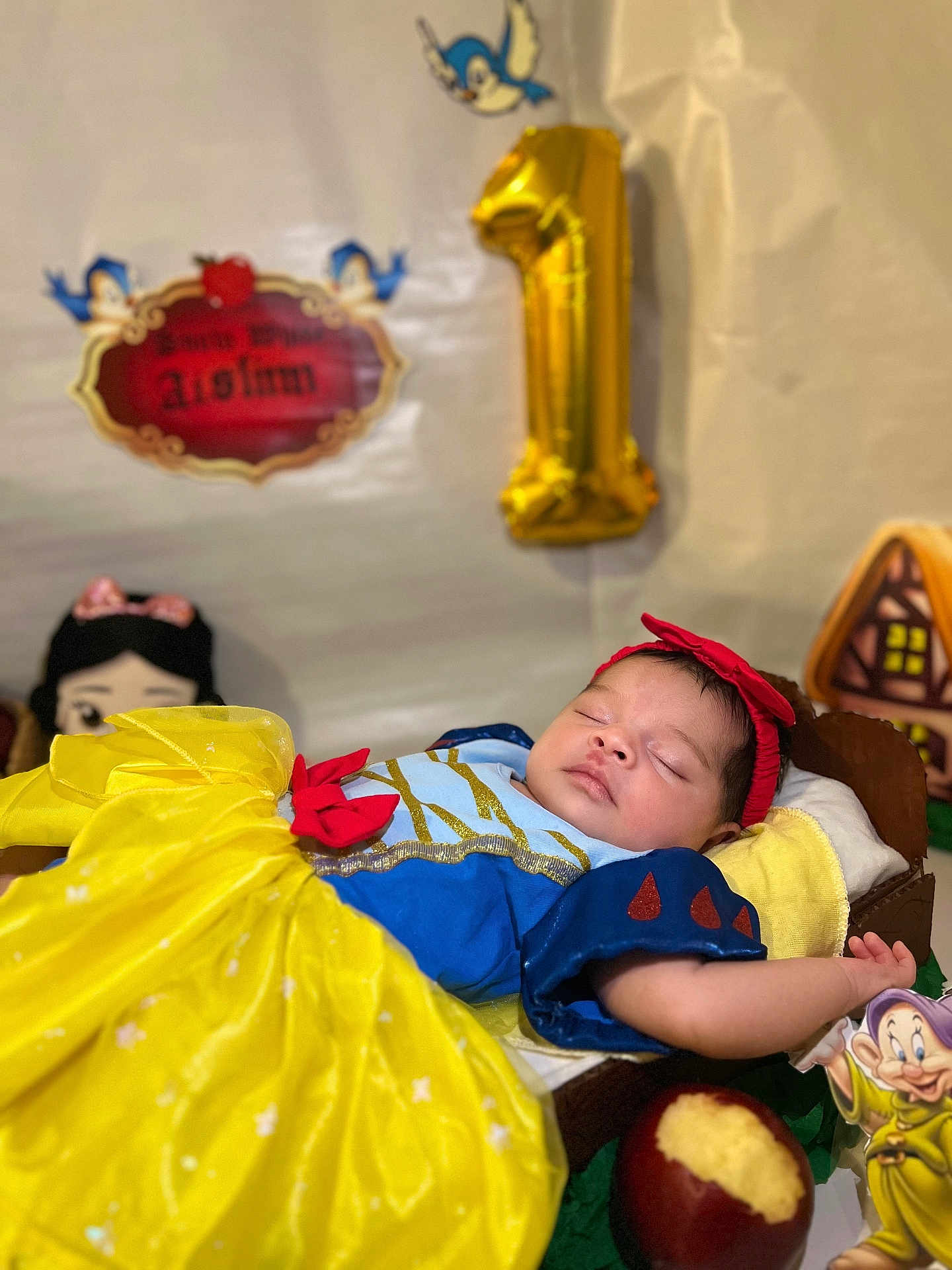 Aislinn is registered to the contest to win money with this photo: baby, sleeping, snow_white_costume, yellow_dress, red_headband, pillow, decorations, gold_balloon, number_one, bitten_apple, dwarf_figurine, fairy_tale, birthday, costume, cute, portrait, child, celebration, indoor, soft_lighting