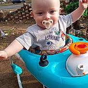 Justen is registered to the contest to win money with this photo: baby, pacifier, walker, outdoor, child, toddler, shirt, astronaut, toy, blue, happy, reaching, face, person, daylight, car, grass, tree, brick_wall, shoe