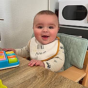 Malo a rejoint le concours — aidez-le/la à gagner de superbes lots ! baby, child, high_chair, bib, toy, table, wood, smiling, happy, indoor, face, person, clothing, kitchen, furniture, playing, cute, infant, seat, expression