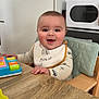 baby, child, high_chair, bib, toy, table, wood, smiling, happy, indoor, face, person, clothing, kitchen, furniture, playing, cute, infant, seat, expression