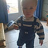 child, toddler, indoor, cookie, striped_clothing, overalls, blue_shoes, door, sunlight, curtain, tile_floor, furniture, box, window, person, young_child, standing, holding, casual_clothing, home