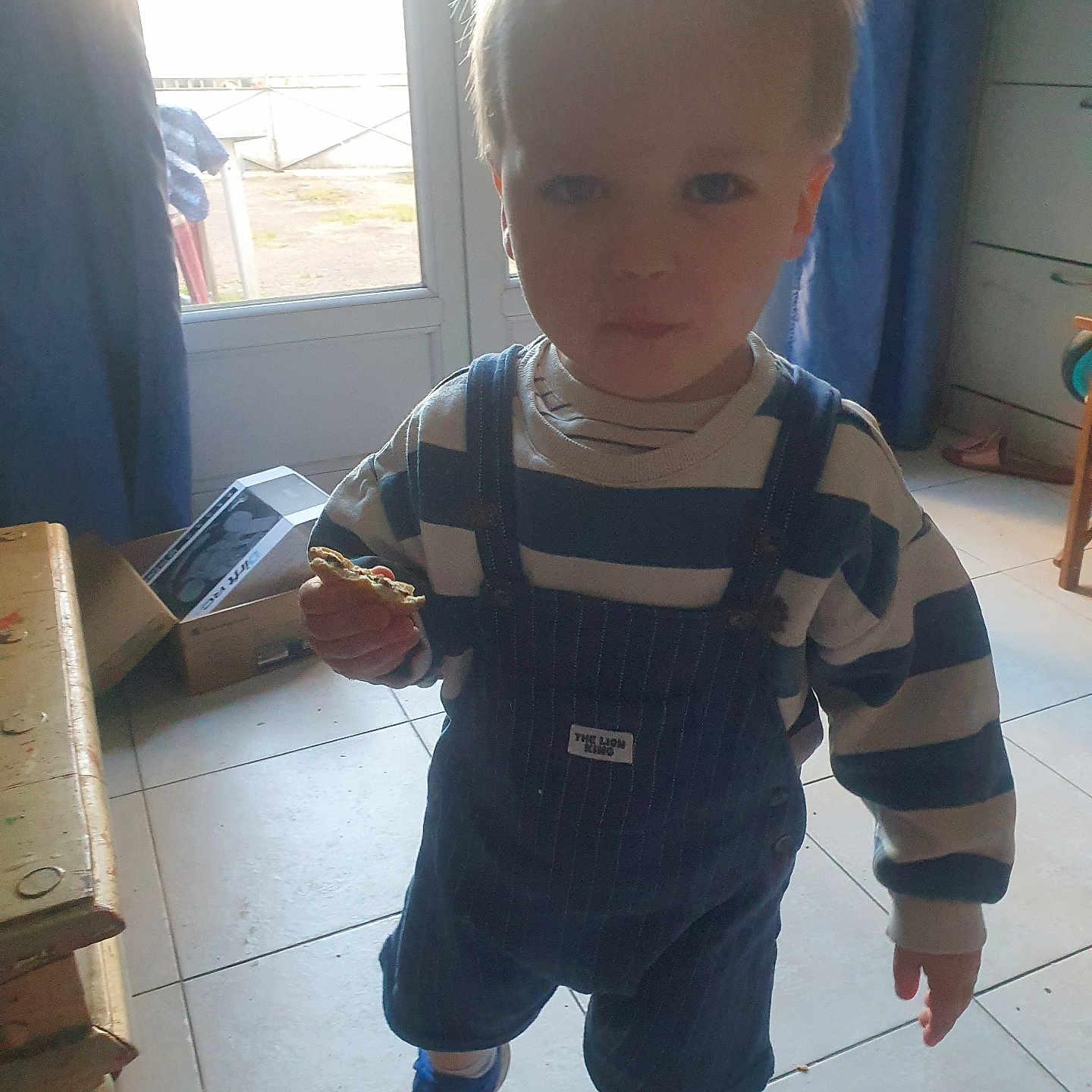 Timéo a rejoint le concours — aidez-le/la à gagner de superbes lots ! child, toddler, indoor, cookie, striped_clothing, overalls, blue_shoes, door, sunlight, curtain, tile_floor, furniture, box, window, person, young_child, standing, holding, casual_clothing, home
