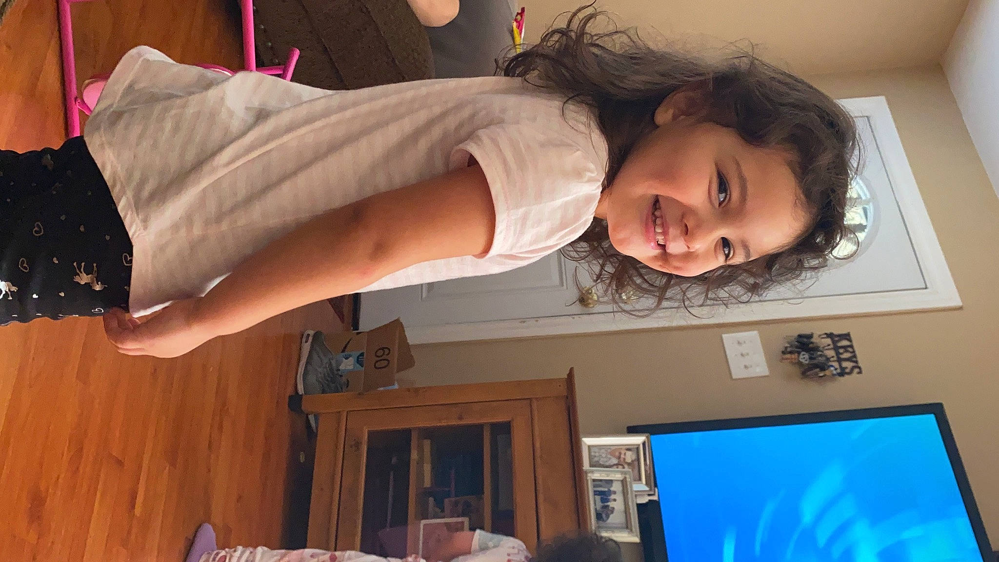 Emma is registered to the contest to win money with this photo: computer_monitor, display_device, dress, elbow, fun, happy, hardwood, human_leg, joint, joy, person, room, smile, standing, television, television_set, thigh, toddler, trunk, waist