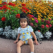 Mandaara is registered to the contest to win money with this photo: toddler, child, flower_bed, flowers, outdoor, plants, greenery, curb, sidewalk, romper, sneakers, hair_tied_up, yellow_flowers, red_flowers, pink_flowers, white_flowers, daylight, nature, casual_clothing, garden