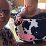 Emrick is registered to the contest to win money with this photo: baby, child, adult, glasses, plush, cow_pattern, basket, indoor, curious, smile, person, face, table, clothing, short_sleeves, hand, light, background, blur, portrait