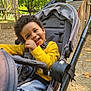 toddler, child, stroller, yellow_shirt, smiling, happy, outdoor, park, playground, trees, daylight, curly_hair, seatbelt, casual_clothing, baby_walker, nature, baby, seat, play_area, joyful