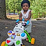 Ky-Mani participe au concours pour gagner de l'argent avec cette photo : toddler, child, toy_walker, outdoor, trees, greenery, pavement, fence, curly_hair, summer_clothing, play, sunlight, nature, park, smiling, walking, colorful_toy, plastic_toy, daytime, young_child