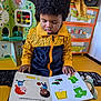 Ky-Mani a rejoint le concours — aidez-le/la à gagner de superbes lots ! child, toddler, book, reading, curly_hair, yellow_jacket, navy_jacket, toy, furniture, drawer, room, indoor, colorful, playroom, floor_mat, hand, face, socks, illustration, education