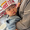 Rocky joined the competition — help win amazing prizes! baby, child, infant, face, person, hat, paper_hat, clothing, denim_jacket, adult, arm, hand, indoor, restaurant, chair, table, floor, red_chair, casual, holding