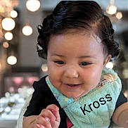 Kross is registered to the contest to win money with this photo: baby, child, smile, curly_hair, bib, clothing, face, hand, indoor, bokeh, lighting, person, cute, portrait, happy, infant, closeup, skin, young, expression