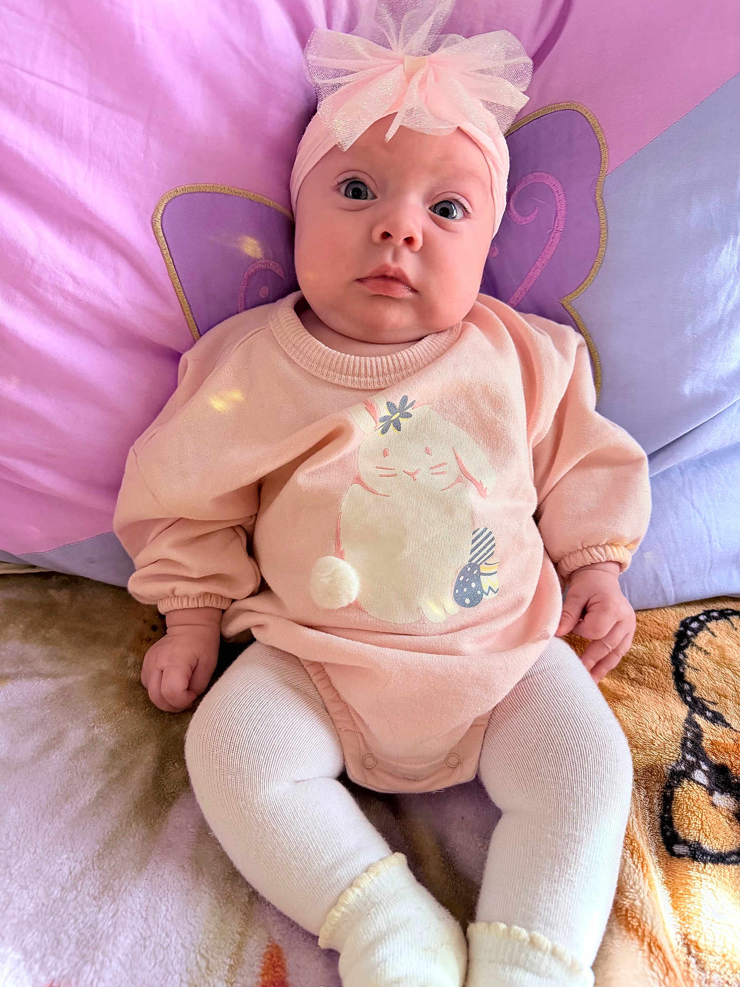 Aylani joined the competition — help win amazing prizes! baby, infant, headband, bow, pink_clothing, sweater, bunny_design, pom_pom, blanket, cushion, purple_background, seated, cute, wide_eyes, soft_lighting, indoors, child, toddler, sock, portrait