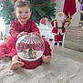 bare_feet, big_eyes, child, christmas_tree, festive, fur_rug, gift_box, holiday_decor, indoor, lace_dress, large_bow, ornament, red_dress, santa_figure, sitting, smile, snow_globe, toddler, tree_decorations, wooden_floor