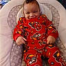 baby, child, pajamas, red_clothing, smiling, face, cushion, chair, indoor, curly_hair, comfort, happy, infant, person, patterned_clothing, folded_blankets, home, seated, cute, toddler