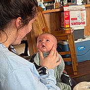 Waylon George is registered to the contest to win money with this photo: baby, woman, indoor, kitchen, holding, blanket, wooden_chair, cabinet, diapers, drink, table, person, face, child, curious, clothing, furniture, light, home, portrait