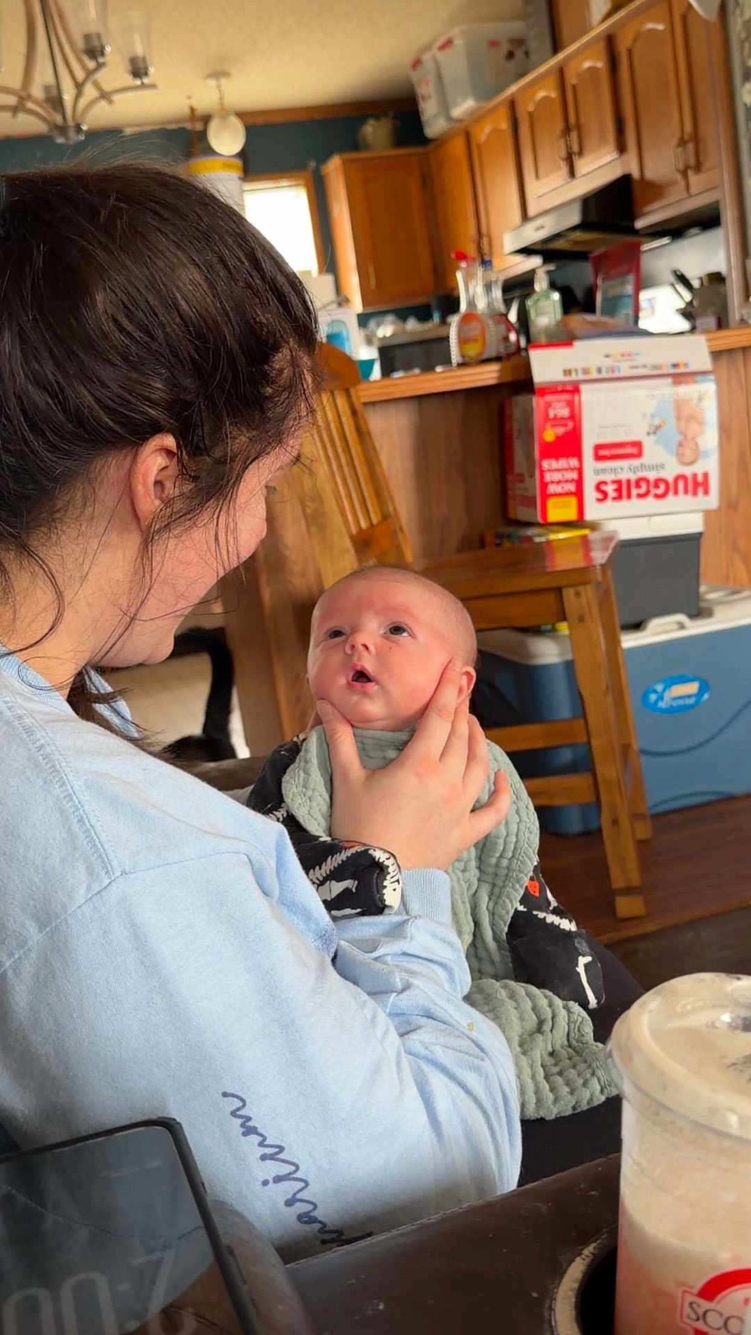 Waylon George is registered to the contest to win money with this photo: baby, woman, indoor, kitchen, holding, blanket, wooden_chair, cabinet, diapers, drink, table, person, face, child, curious, clothing, furniture, light, home, portrait