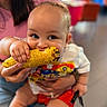 baby, child, corn, food, eating, hand, person, clothing, red_shorts, white_shirt, indoor, blurred_background, adult_hand, grilled_corn, smiling, cute, closeup, portrait, messy_eating, joy