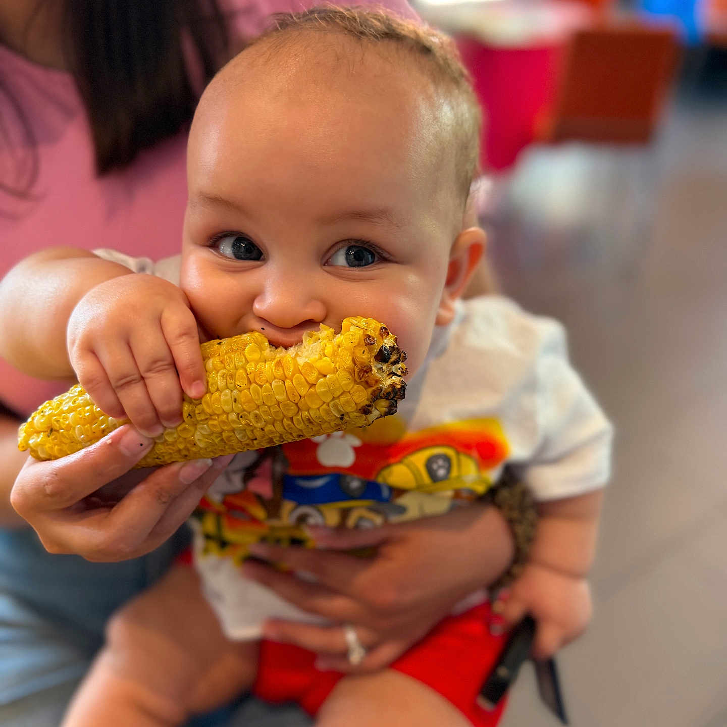 Serenity is registered to the contest to win money with this photo: baby, child, corn, food, eating, hand, person, clothing, red_shorts, white_shirt, indoor, blurred_background, adult_hand, grilled_corn, smiling, cute, closeup, portrait, messy_eating, joy