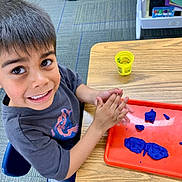 Ezekiel is registered to the contest to win money with this photo: child, boy, playdough, red_tray, table, classroom, smiling, short_hair, blue_playdough, yellow_container, carpet, chair, wooden_table, indoor, happy, hands, toy, education, preschool, activity