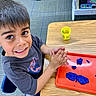 activity, blue_playdough, boy, carpet, chair, child, classroom, education, hands, happy, indoor, playdough, preschool, red_tray, short_hair, smiling, table, toy, wooden_table, yellow_container