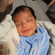 Ezra is registered to the contest to win money with this photo: baby, smiling, sleeping, blanket, blue_onesie, infant, cozy, indoor, cute, child, portrait, newborn, warm_light, soft_texture, peaceful, happy, face, skin, head, person