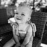 toddler, child, black_and_white, smiling, sitting, wicker_chair, curly_hair, outdoor, portrait, happy, baby_clothes, small_hands, cute, person, one_person, daylight, seat, photography, expression, innocence
