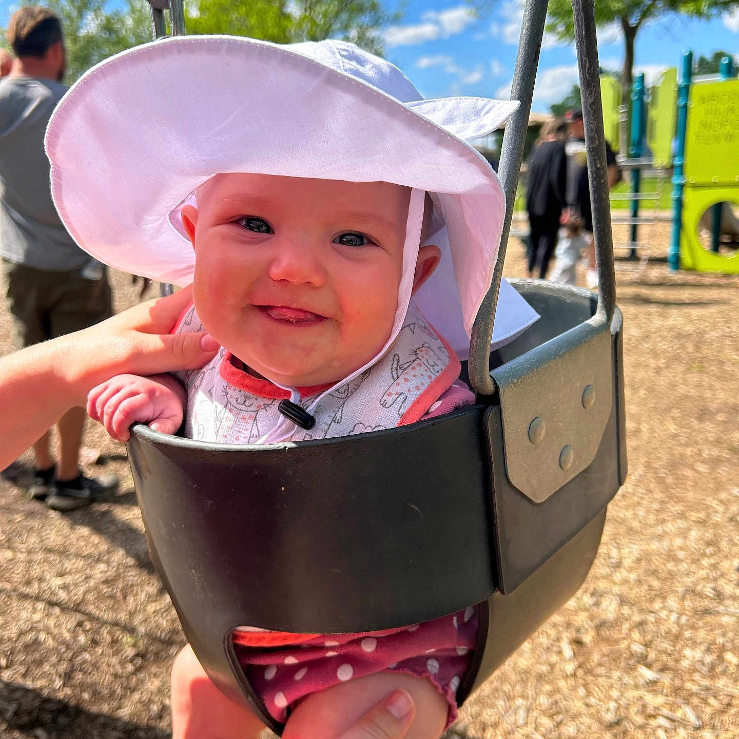 Mackenzie is registered to the contest to win money with this photo: adult, baby, blurred_background, child, daylight, hands, happy, hat, holding, metal, outdoor, person, playground, seat, smiling, sun_hat, swing, tongue_out, trees, wood_chips