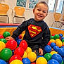 Liam a rejoint le concours — aidez-le/la à gagner de superbes lots ! toddler, child, smiling, superman_logo, ball_pit, colorful_balls, indoor, playroom, wooden_chairs, windows, natural_light, happy, casual_clothing, playful, person, young_child, room, daylight, fun, seated