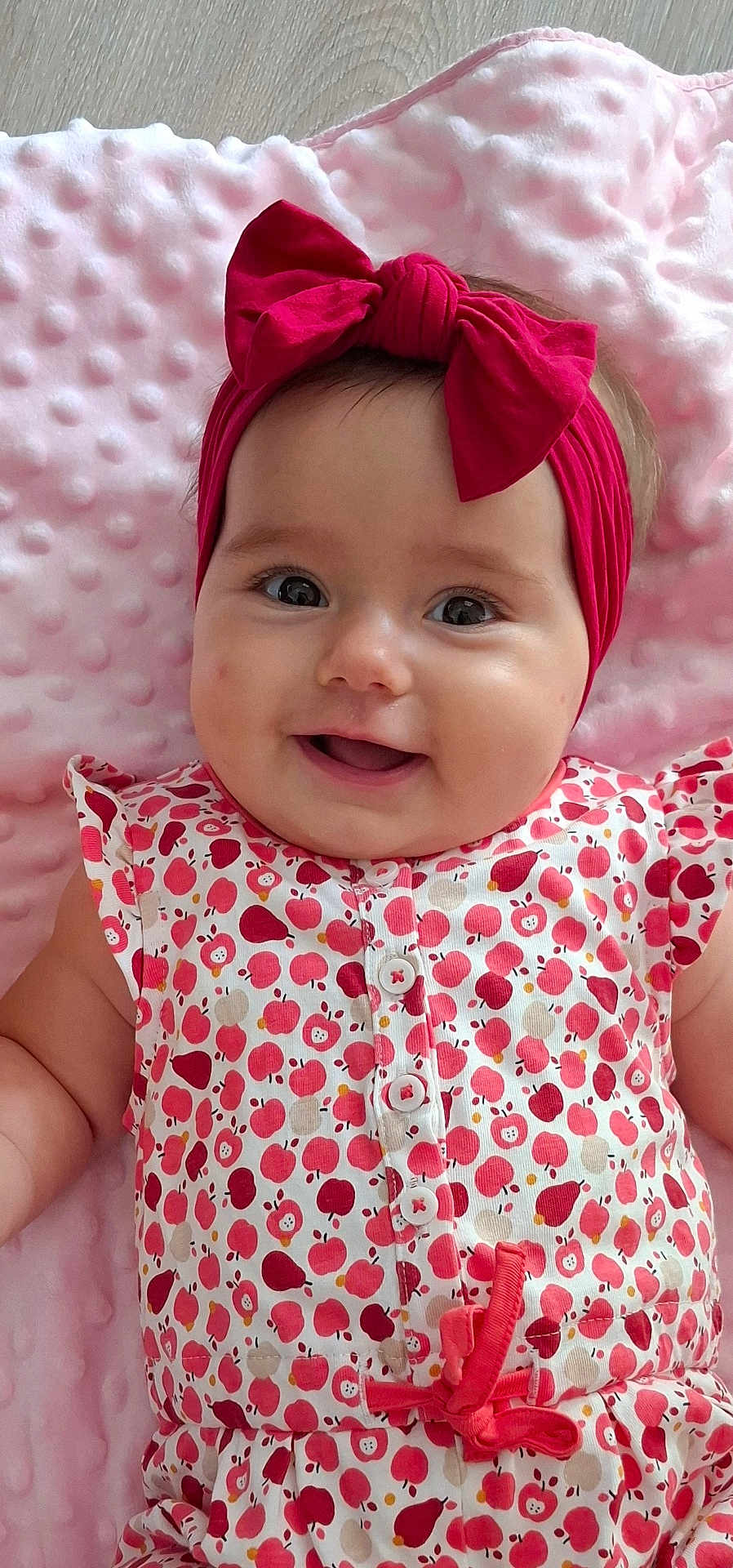 Juliette a rejoint le concours — aidez-le/la à gagner de superbes lots ! baby, infant, smile, headband, red_bow, cute, patterned_clothing, apple_pattern, pink_blanket, happy, portrait, child, closeup, soft_texture, lying_down, adorable, young_child, face, eyes, buttoned_outfit