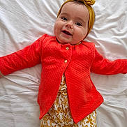 Juliette participe au concours pour gagner de l'argent avec cette photo : baby, smiling, child, orange_jacket, headband, bow, white_blanket, floral_pants, lying_down, happy, infant, cute, portrait, indoors, clothing, face, person, young_child, headwear, bright_colors
