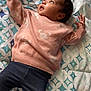 baby, infant, child, sweater, pants, sock, bed, blanket, pattern, lying_down, reaching_hand, curious, indoor, cute, portrait, face, head, skin, person, young