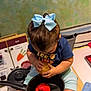 toddler, child, bow, hair_accessory, toy_potato, red_bell_pepper, pot, kitchen_counter, recipe_book, socks, blue_shirt, playing, indoor, cooking_play, pretend_play, curious, baby, seat, table, hand