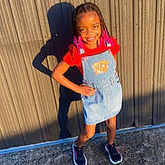 Robyn is registered to the contest to win money with this photo: child, girl, smiling, outdoor, sunlight, shadow, denim_dress, red_shirt, braided_hair, pink_beads, sneakers, concrete_ground, wall, standing, posing, happy, casual_clothing, daylight, fashion, portrait