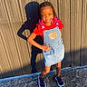 child, girl, smiling, outdoor, sunlight, shadow, denim_dress, red_shirt, braided_hair, pink_beads, sneakers, concrete_ground, wall, standing, posing, happy, casual_clothing, daylight, fashion, portrait