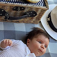 Dylan is registered to the contest to win money with this photo: baby, infant, white_outfit, blanket, picnic_basket, bananas, grapes, dessert, candle, number_two, hat, airplane_pin, checkered_pattern, food, fruit, lying_down, curious, indoors, closeup, portrait