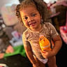 child, toddler, smiling, curly_hair, holding_object, sippy_cup, indoor, toy, furniture, blurred_background, person, happy, standing, casual_clothing, young_child, playroom, home, cute, portrait, warm_lighting