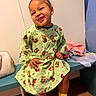 child, toddler, smiling, sitting, hospital_gown, yellow_socks, earring, bench, indoor, clothing, happy, face, person, wooden_floor, bag, curtain, wall, casual, cute, portrait