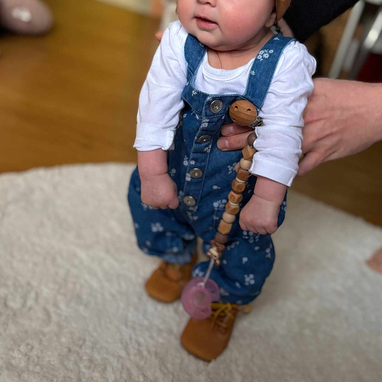 June a rejoint le concours — aidez-le/la à gagner de superbes lots ! baby, blue_overalls, brown_shoes, carpet, child, curious_expression, cute, footwear, hands, headband, home, indoor, infant, pacifier, person, soft_rug, standing, support, toy_clip, wooden_floor