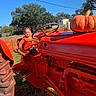Alani is registered to the contest to win money with this photo: toddler, child, tractor, pumpkin, orange_clothing, smiling, outdoor, daylight, adult, vehicle, farm, autumn, grass, tree, blue_sky, fence, person, happy, steering_wheel, celebration