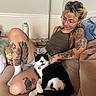 woman, cat, tattoo, glasses, sitting, couch, indoor, petting, black_and_white_cat, relaxed, shorts, tank_top, plush_toy, water_bottle, tattooed_arm, tattooed_leg, casual, home, comfort, smile