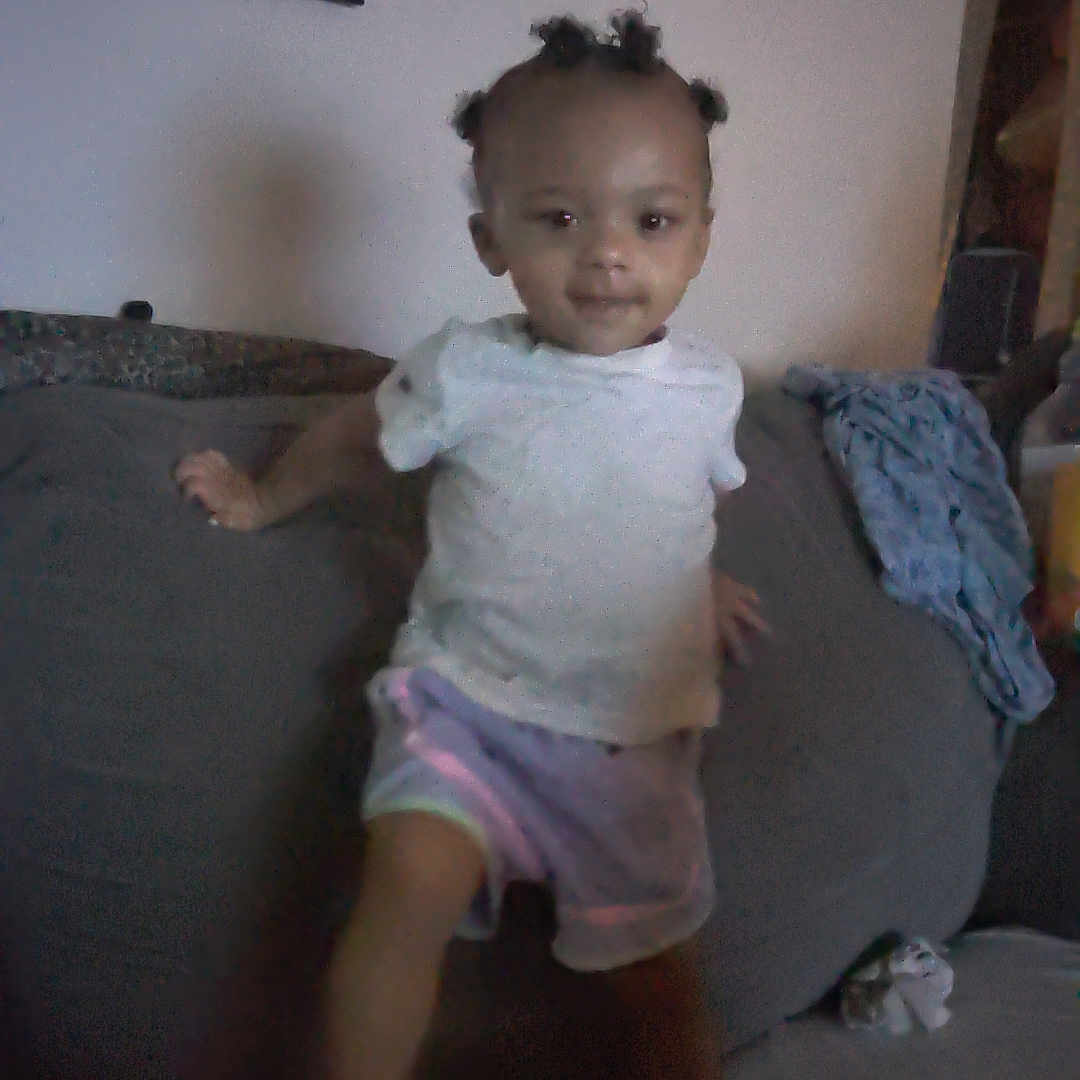 Toshia Harris is registered to the contest to win money with this photo: blanket, casual_clothing, child, couch, curious, dim_light, furniture, hair, home, indoor, living_room, person, portrait, shorts, smiling, standing, toy, wall_art, white_shirt, young_child