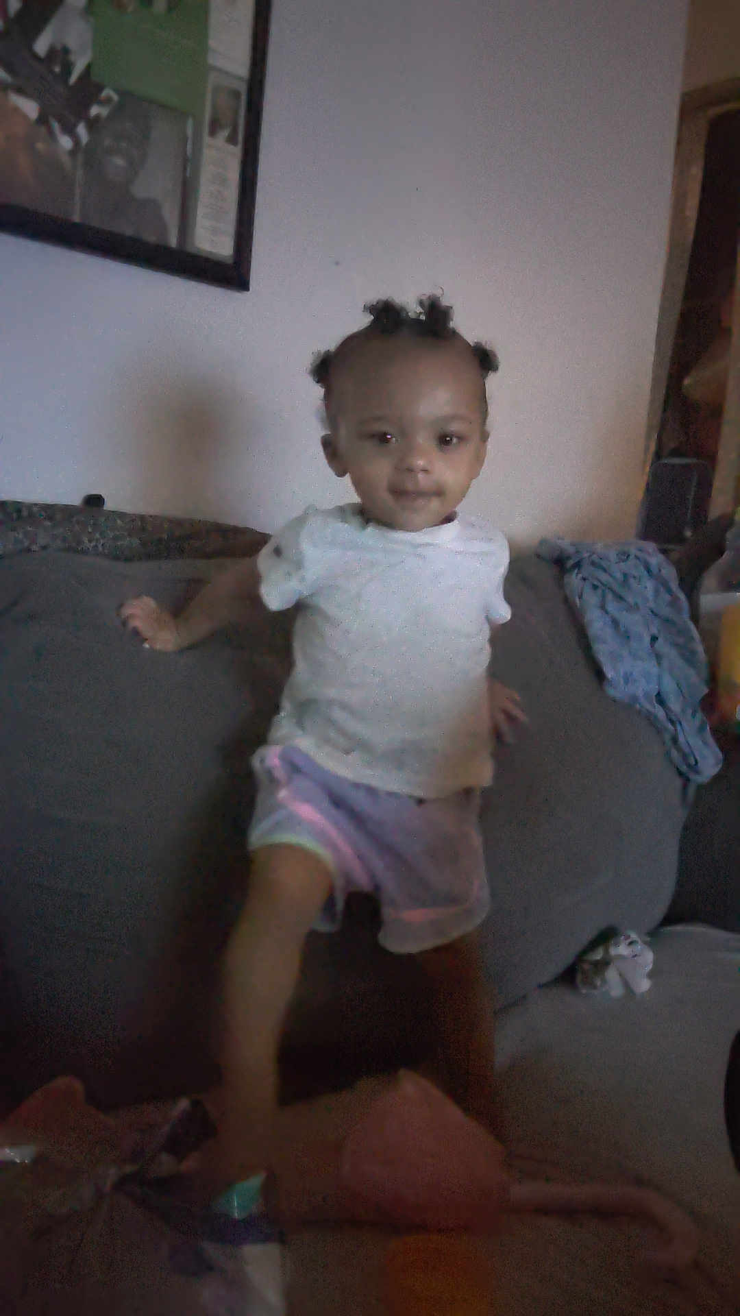 Toshia Harris is registered to the contest to win money with this photo: child, indoor, couch, person, hair, white_shirt, shorts, toy, blanket, wall_art, furniture, home, living_room, young_child, curious, standing, casual_clothing, dim_light, portrait, smiling