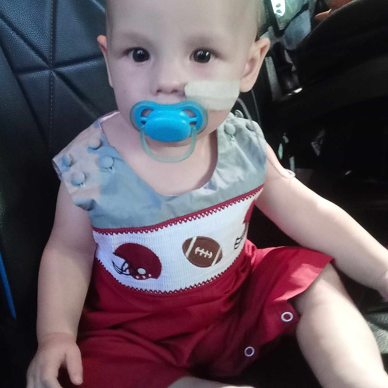 Kyland is registered to the contest to win money with this photo: baby, car_seat, child, clothing, cute, face, foot, football_theme, hand, head, indoor, infant, medical_bandage, pacifier, person, red_outfit, seat, sitting, transport, young_child