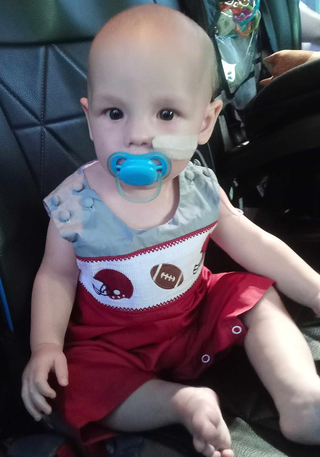 Kyland is registered to the contest to win money with this photo: baby, child, pacifier, car_seat, medical_bandage, red_outfit, football_theme, sitting, indoor, cute, face, head, hand, foot, person, clothing, infant, young_child, seat, transport