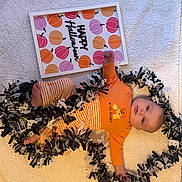 Aiyanna is registered to the contest to win money with this photo: baby, infant, halloween, orange_clothing, striped_pants, blanket, garland, ghosts, pumpkins, sign, holiday, festive, cute, lying_down, indoors, decorations, child, celebration, smiling, portrait